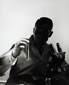 Science And Technology Gallery: Male scientist wearing lab coat, sitting in darkness behind microscope