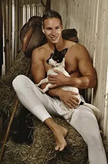 Desire Gallery: Man with a naked torso wearing long underwear with a dog in a horse barn sitting on bales of hay