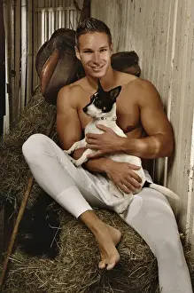 Blond Hair Prints: Man with a naked torso wearing long underwear with a dog in a horse barn sitting on bales of hay