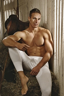 Desire Gallery: Man with a naked torso wearing long underwear in a horse barn sitting on bales of hay
