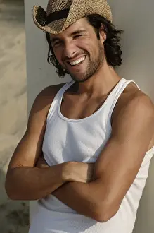 Man wearing a sun hat on a beach, leaning against a cottage, smiling