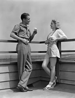 Man and woman smiling at each other, standing outside leaning against railing