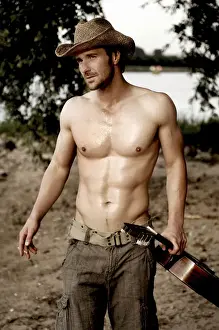 Musical Instrument Prints: Masculine man without a shirt, wearing a cowboy hat and holding a guitar on a beach