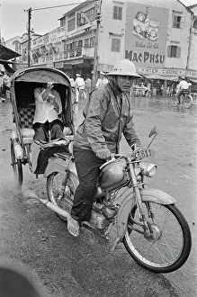 Prints of A motorcycle rickshaw driving through the rain, the passenger sheltered by a canopy, and the driver wearing a safari-style hat, in Saigon, Vietnam, 30th August 1965 Vietnam Prints: Motorcycle Rickshaw, Saigon