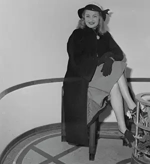 Nancy Guild Wearing Hat and Coat and Sitting on a Stool