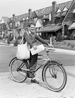 Images Dated 30th June 2008: Paperboy on bicycle in suburban neighborhood, with sack over shoulder and folded newspaper in hand