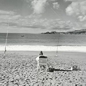 Leisure Collection: Person sitting on beach with fishing poles embedded in sand and using cell phone