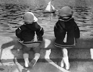 Twins Prints: Twin girls (6-7) looking at model sailboat in pond