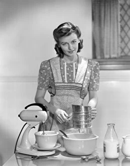 Preparation Gallery: Woman In A Apron Over A Cotton Print Dress Sifting Flour In A Bowl Between A Mixer & A