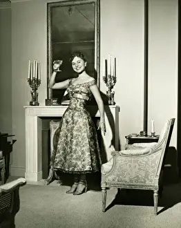 Woman in evening gown toasting by fireplace, (B&W), portrait