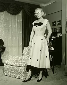 35 39 Years Collection: Woman in fashionable dress standing by armchair in living room, (B&W), portrait