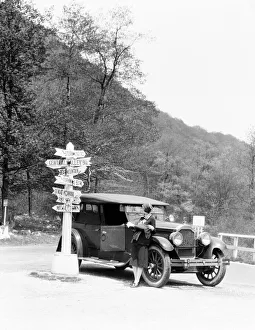Driver Gallery: Woman leaning on car looking at road map and signs with arrows pointing to selected towns