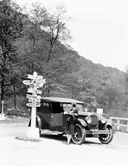 One Mid Adult Woman Only Prints: Woman leaning on car looking at road map and signs with arrows pointing to selected towns