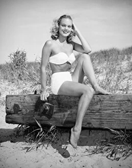 Iconic Bikini Collection: Woman wearing bikini sitting on log on beach, (B&W), (Portrait)