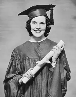 Woman wearing graduation gown holding diploma, (B&W), portrait