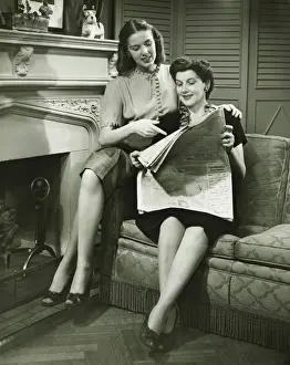 Two women sitting by fireplace, reading newspaper, (B&W)