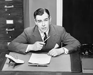 Calendar Prints: Young businessman sitting at small desk, holding pencil, (B&W)