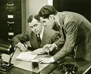 Bending Down Prints: Two young businessmen talking at small desk, (B&W)