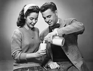 20 25 Years Gallery: Young couple pouring milk from bottle to glass in studio, (B&W)