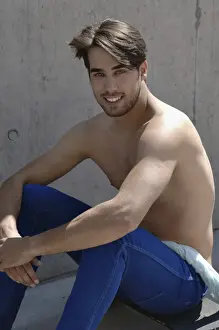 Young shirtless skater in front of a concrete wall, sitting on his skateboard