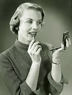 Permed Hair Prints: Young woman applying lipstick, looking at powder compact mirror, (B&W), close-up