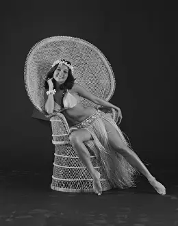 1980 Prints: Young woman hula dancer in traditional clothing sitting on wicker chair, smiling, portrait