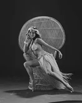 1980 Prints: Young woman hula dancer in traditional clothing sitting on wicker chair, smiling, portrait