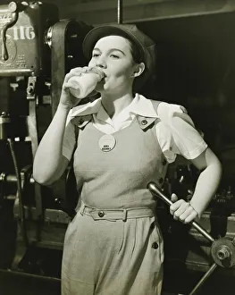 Bib Overalls Gallery: Young woman in overalls standing by lathe in factory, drinking milk, (B&W)