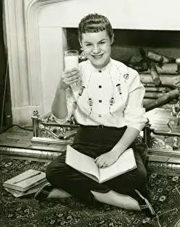 Carpet Prints: Young woman sitting by fireplace, holding glass of milk in raised hand, (B&W), portrait